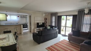 Una sala de estar moderna con un sofá negro, un sofá marrón y una mesa de comedor con sillas. Hay una zona de cocina con televisor, refrigerador y ventana con cortinas.