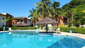 Una piscina con sillas de descanso y sombrillas de paja, rodeada de palmeras y un edificio con antenas parabólicas.