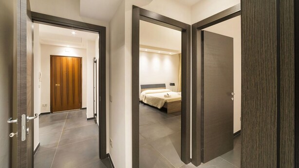 Un pasillo de hotel con una puerta de madera al final, que da a un dormitorio con una cama y una mesita de noche.