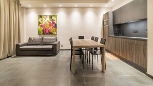Una sala de estar moderna con una mesa de comedor y sillas, un sofá, una zona de cocina con armarios y una encimera, y un cuadro grande con motivos florales en la pared.