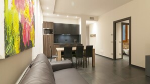 Una sala moderna con un sofá gris, un comedor con una mesa y sillas de madera, una cocina con armarios oscuros y un televisor grande, y un baño visible a través de una puerta abierta.