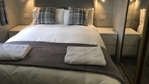 Una cama bien tendida con sábanas blancas y almohadones a cuadros, una mesita de noche con un control remoto y un espejo en la pared.