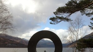 Un perro frente a una escultura circular junto a un lago con montañas al fondo.