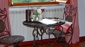 Une pièce avec vue sur les montagnes, visible à travers une fenêtre ornée de rideaux roses. On y trouve une table avec une cafetière et un vase rempli de fleurs. Deux chaises sont placées autour de la table.