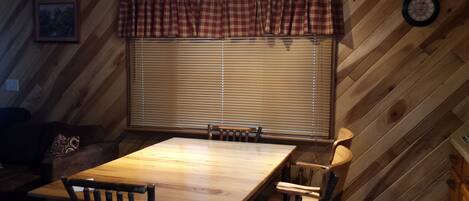 A dining area with a wooden table and chairs, a window with blinds, a plaid valance, and a clock on the wall.