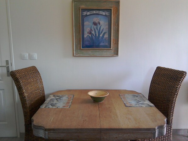 A dining table with two wicker chairs, a bowl, and a framed picture of iris plants on the wall. - Perfect hideaway in a rural setting, yet only a 10 minute walk to the village. (Cerisy-La-Forêt)