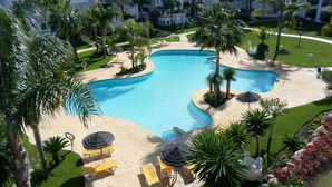 En pool på semesteranläggningen, omgiven av palmer, med solstolar och utsikt över hotellbyggnaderna.