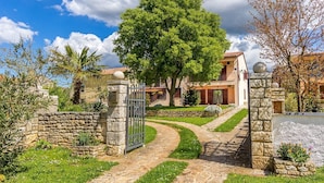 A stone-walled property with a gated entrance, a large tree, and a pathway leading to a building with a tiled roof. - Apartment with large garden to play football (Gajana)