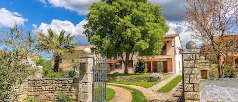 A stone-walled property with a gated entrance, a large tree, and a pathway leading to a building with a tiled roof.