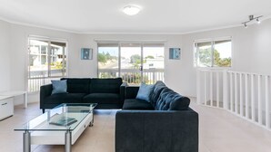 A modern living room with a dark grey sectional sofa, a glass coffee table, and a white cabinet. Large windows offer views of the outside.