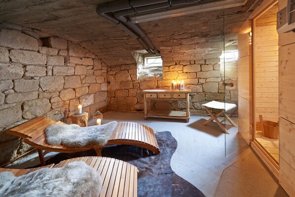 A rustic stone-walled room with wooden furniture, a fur-covered bench, and a small table with candles.