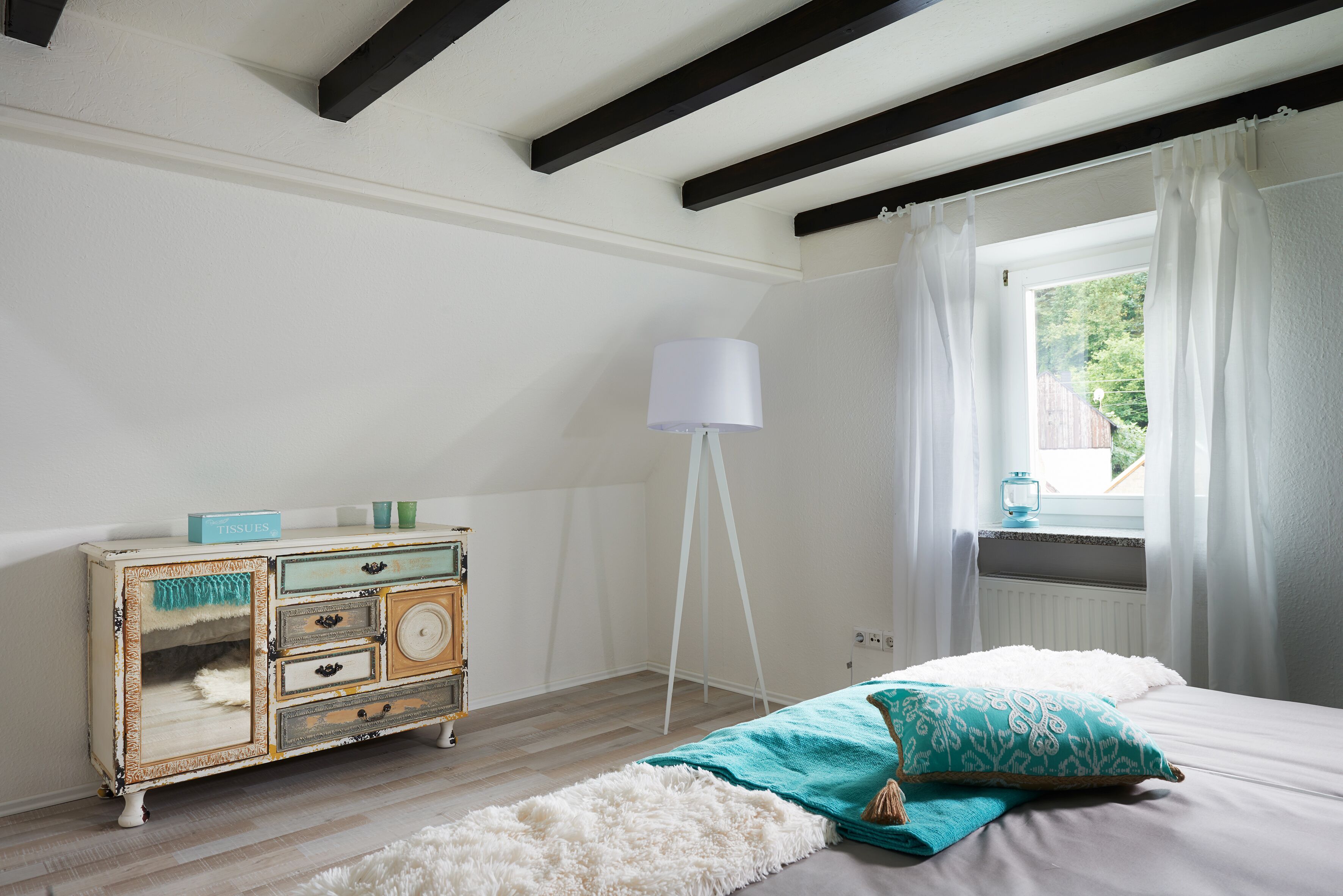 Une chambre avec une commode ancienne, une lampe blanche, un tapis moelleux et une fenêtre avec des rideaux.