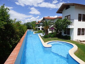 A swimming pool with a curved edge, surrounded by greenery and a white building with a red roof.