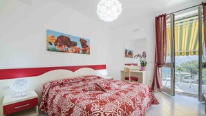 Un dormitorio con cama, escritorio, silla, planta en maceta y balcón con vistas.