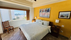 Habitación de hotel con una cama grande, una silla de madera, una mesita de noche y un cuadro de palmeras.