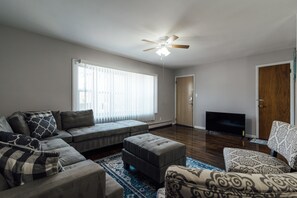 A living room with a sectional sofa, ottoman, patterned armchair, ceiling fan, and a flat-screen TV. - 3 Bedroom near Journal Square (Jersey City)