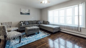 A living room with a sectional sofa, patterned armchair, and ottoman, a ceiling fan, and a large window with blinds.