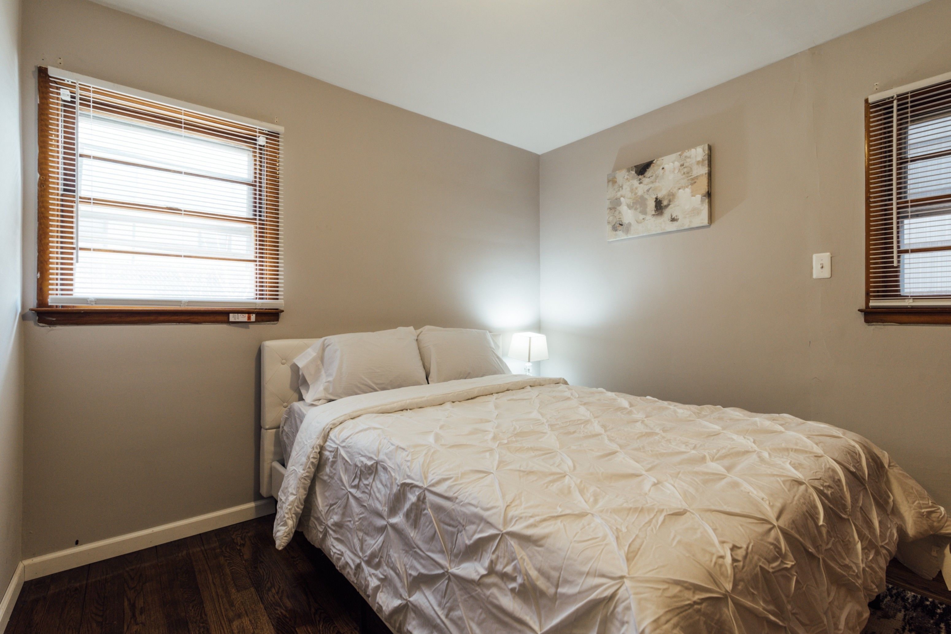 A bedroom with a bed, two windows with blinds, a wall-mounted light, and a small painting.