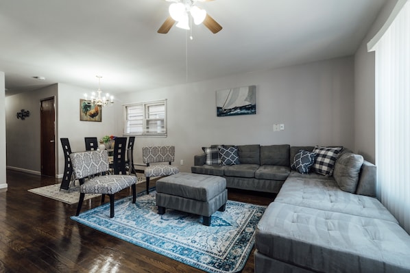 A living room with a gray sectional sofa, a gray ottoman, patterned chairs, a chandelier, and a ceiling fan. - 3 Bedroom near Journal Square (Jersey City)