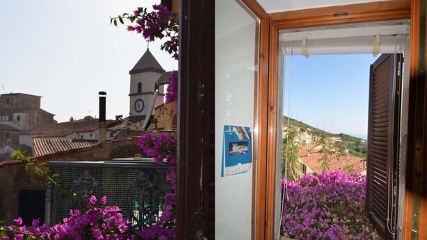 A view from a balcony with purple flowers, a clock tower, and a building with a tiled roof. - Il Rifugio - Capoliveri  - IL RIFUGIO (Capoliveri)
