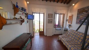 A room with a wooden bench, a bed with a checkered bedspread, a television on a shelf, and a window with a view of greenery. - Il Rifugio - Capoliveri  - IL RIFUGIO (Capoliveri)