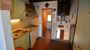 A compact kitchen with a gas stove, sink, and a small refrigerator. A wooden door leads to a bathroom with a toilet and sink. - Il Rifugio - Capoliveri  - IL RIFUGIO (Capoliveri)