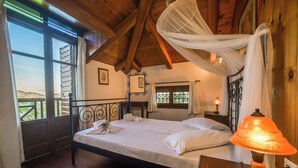 Une chambre avec un plafond en bois, un lit recouvert de draps blancs, une table de chevet avec une lampe, et une vue sur l’extérieur par une fenêtre.