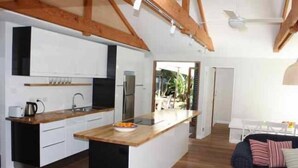 A modern kitchen with white cabinets, a wooden countertop, and a built-in island. There is a dining area with a dark-colored sofa and a white table. - Tui Cottages - House+Cottage - close to beach (Bundeena)