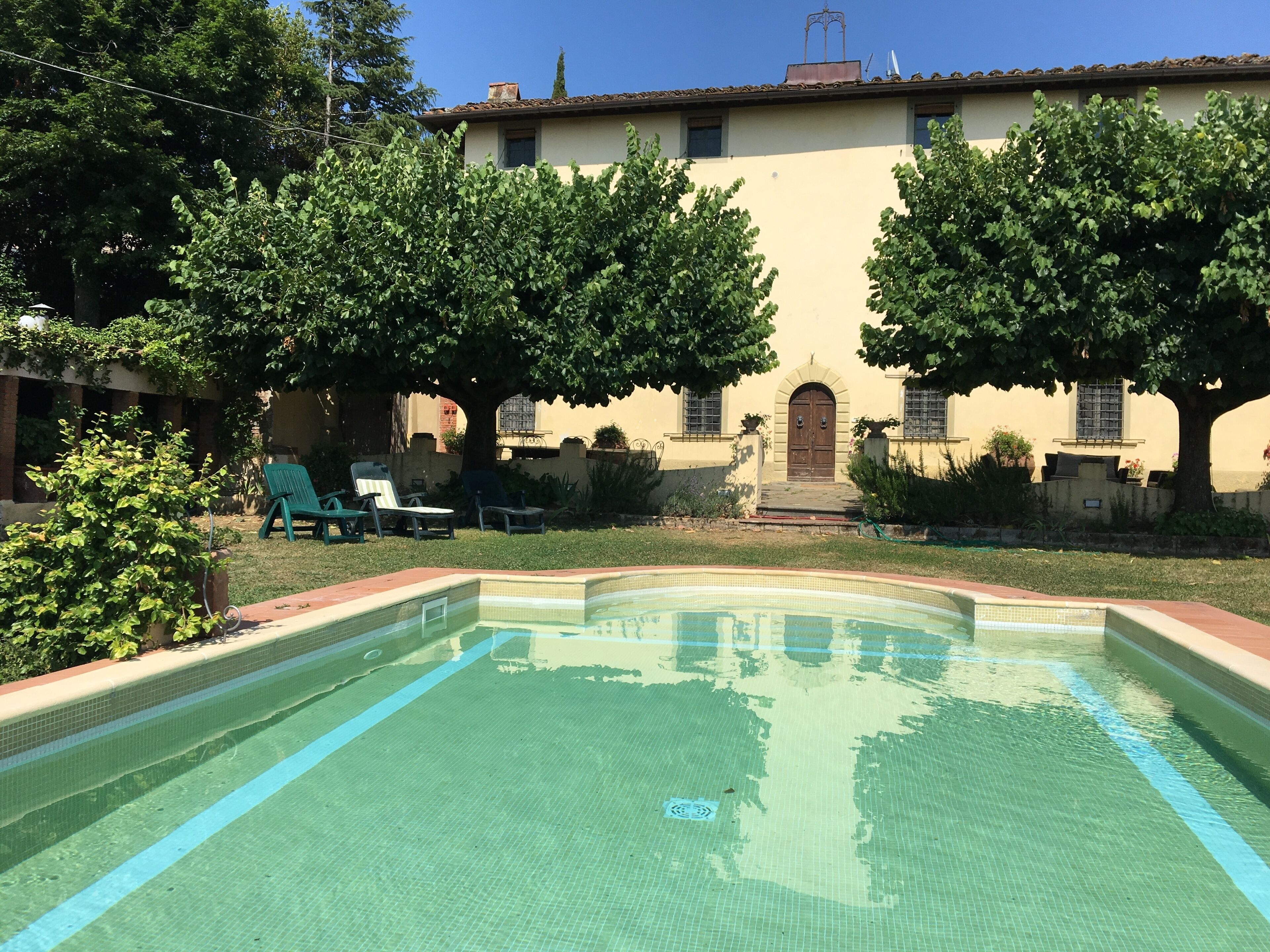 Una piscina con bordes azulejados, rodeada de césped y árboles. Se observan sillas de descanso y un edificio con techo de tejas al fondo.