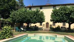 A swimming pool with tiled edges, surrounded by a grassy area and trees. There are lounge chairs and a building with a tiled roof in the background. - Authentic Tuscan Villa with Pool and Panoramic Views near Vinci – Tuscany (lamporecchio)