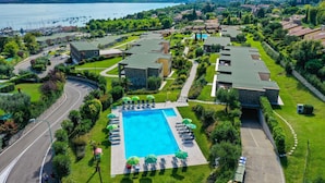 A resort with a large swimming pool surrounded by lounge chairs and umbrellas, set against a backdrop of a lake and residential buildings. - Relais Rosa dei Venti: 1 bedroom elegance, 2 pools, lake view! (Moniga del Garda)