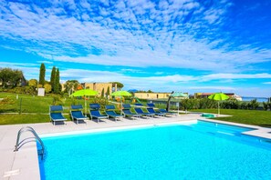 Ein Schwimmbad mit Liegestühlen und Sonnenschirmen, umgeben von Grünflächen und blauem Himmel.