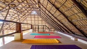 Ein Innenraum mit einem strohgedeckten Dach, Holzboden und farbenfrohen Yogamatten, die in Reihen angeordnet sind.