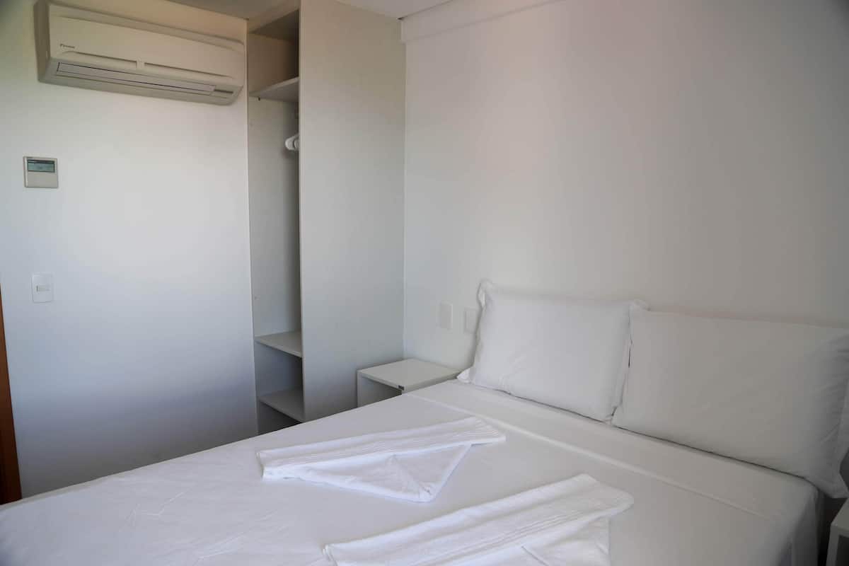 Une chambre d’hôtel avec un lit, du linge de lit blanc, une table de chevet, une petite table et un appareil de climatisation.