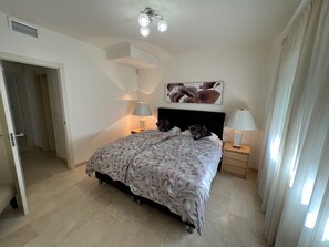 A bedroom with a bed, two bedside tables, a ceiling light fixture, a door, and a wall-mounted artwork.