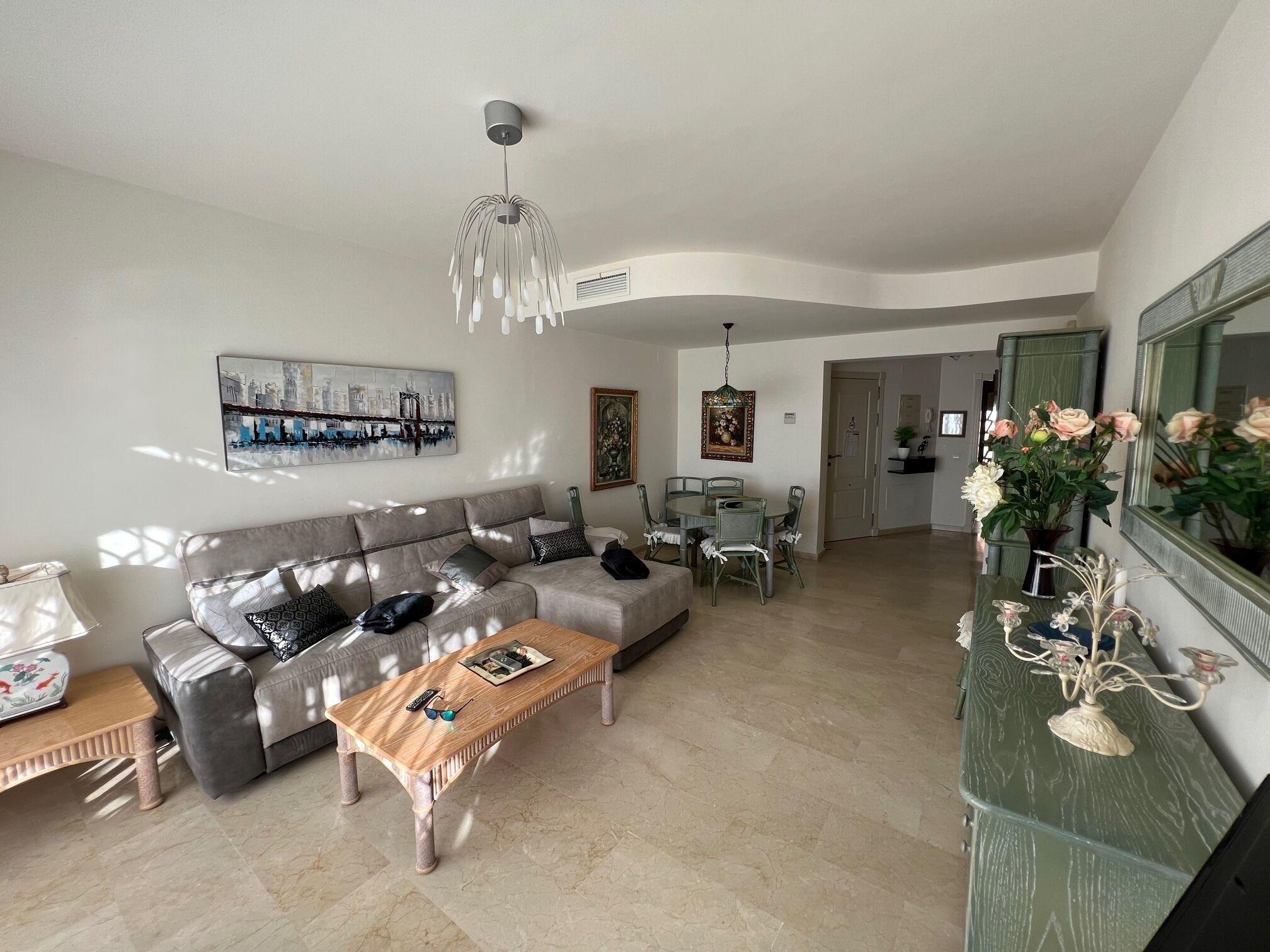 Ein Wohnzimmer mit grauem Sofa, einem Holztisch und einem Esstisch mit Stühlen. An der Wand hängt ein Gemälde und von der Decke ein Kronleuchter.