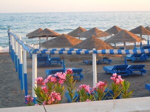 Ein Strand mit blauen Liegestühlen und reetgedeckten Sonnenschirmen, ein sandiger Uferstreifen und rosa Blüten im Vordergrund.