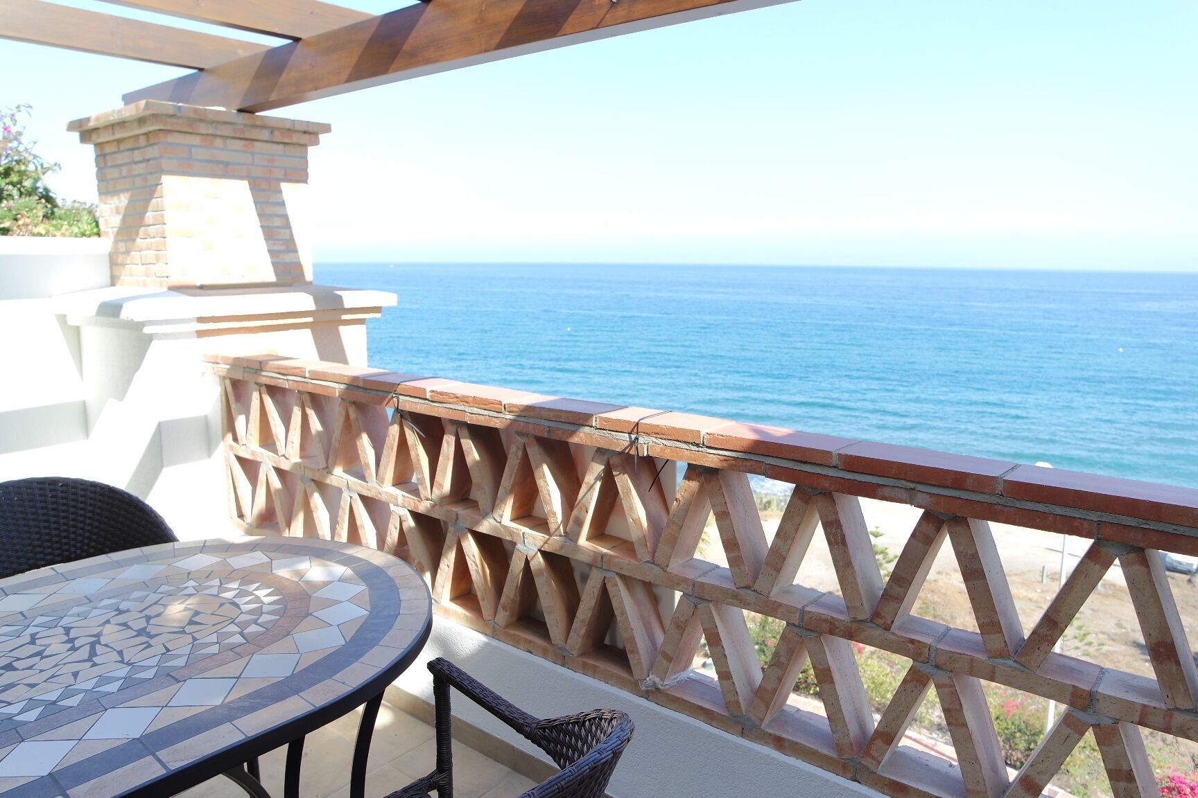 A patio with a mosaic table, wicker chairs, and a wooden railing overlooking the sea.