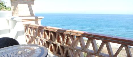 A patio with a mosaic table, wicker chairs, and a wooden railing overlooking the sea.