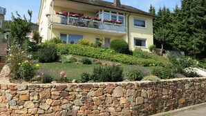 A two-story house with a stone wall in front, a balcony with flowers, and a garden with various plants.
