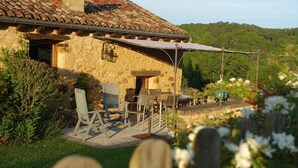 Un bâtiment en pierre avec un toit en tuiles, un espace extérieur aménagé avec des chaises et des tables, et un jardin agrémenté de fleurs et de verdure.