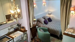 Une chambre d’hôtel compacte comprenant un coin cuisine, un lit avec une literie à motifs, un fauteuil vert et un grand miroir.