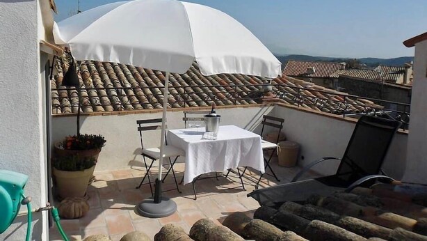 Une terrasse avec une table et des chaises, protégée par un parasol blanc, donnant sur une zone de toiture recouverte de tuiles.