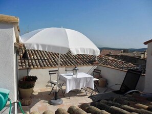 Een terras met een tafel en stoelen onder een witte parasol, met uitzicht op een dak met tegels.