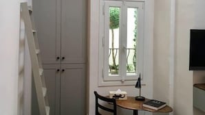 Une petite pièce avec un mur en pierre, une fenêtre donnant sur la verdure, un bureau avec une chaise, un fauteuil vert et un téléviseur fixé au mur.