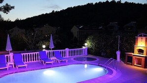 Área de piscina com cadeirões de descanso e churrasqueira, cercada por um muro e árvores.