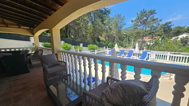 Uma área de estar coberta ao ar livre, com móveis de vime e uma mesa de vidro, com vista para uma piscina e vegetação.