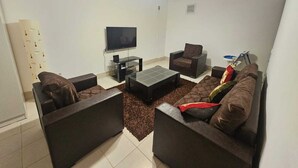 Uma sala de estar com televisão de tela plana, dois poltronas, uma mesa de centro e um ventilador de teto.