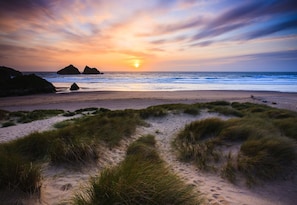 Une plage avec des dunes de sable et de l’herbe au premier plan, un coucher de soleil sur l’océan et des formations rocheuses à l’horizon.
