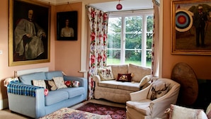 Un salon avec un canapé bleu, un fauteuil beige et un tapis à motifs. Deux tableaux ornent les murs, et une fenêtre est agrémentée de rideaux à fleurs.